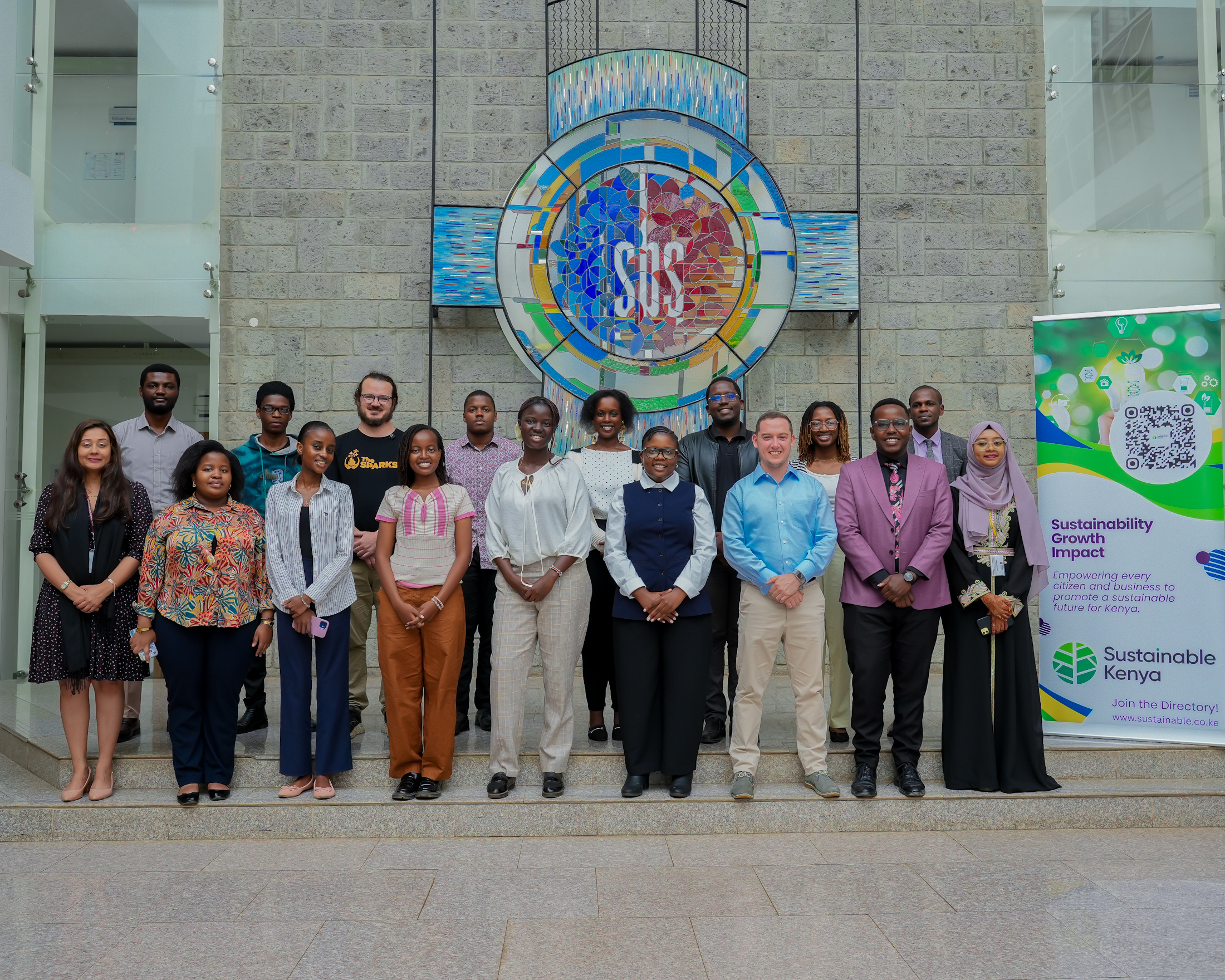Participants of Sustainable Business Internship programme Kenya stand on steps inside a modern building, smiling and facing the camera. Behind them is a large circular stained-glass-style mosaic mounted on a stone wall. On the right side, a standing banner reads “Sustainable Kenya” with messaging about sustainability, growth, and impact.