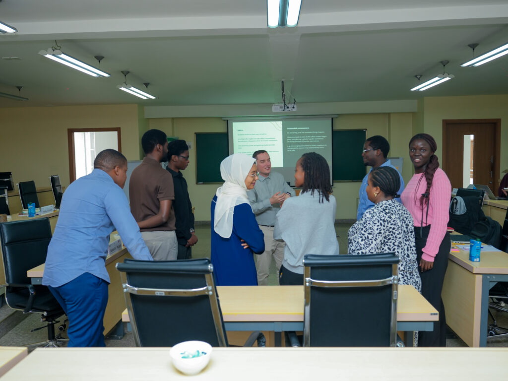 Participants stand in a circle in a classroom-style training room, engaged in a discussion. They wear business-casual attire, including one woman in a white headscarf and blue outfit. Tables with chairs, notebooks, and drinks surround them. At the front, a projector screen displays a slide, with chalkboards on either side. The setting suggests an interactive workshop or group activity.