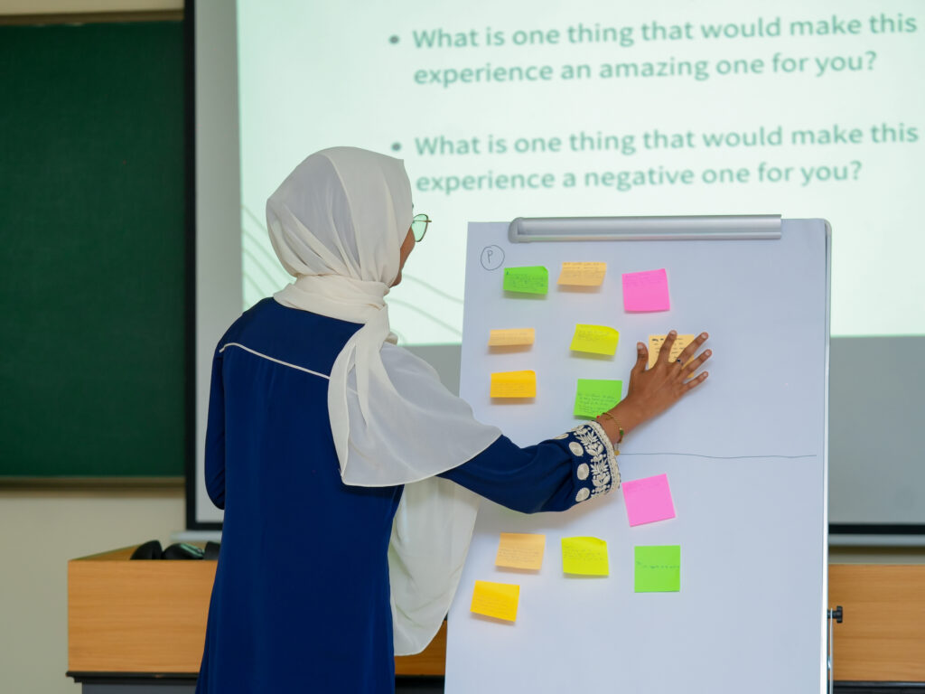 A woman wearing a white headscarf and glasses is seen from behind as she places and organizes colorful sticky notes on a flipchart. The notes are arranged in sections, suggesting a group activity or feedback exercise. In the background, a projected slide shows discussion prompts asking participants what would make an experience positive or negative, indicating an interactive workshop or training session.