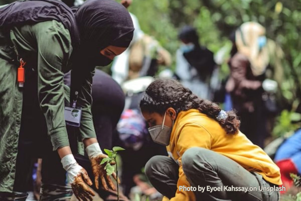 IDGs Blog – Youth Voices in Climate Resilience A woman in a hijab and a girl in a yellow hoodie and face mask kneel in a forest, working together to plant a small green sapling in the soil.
