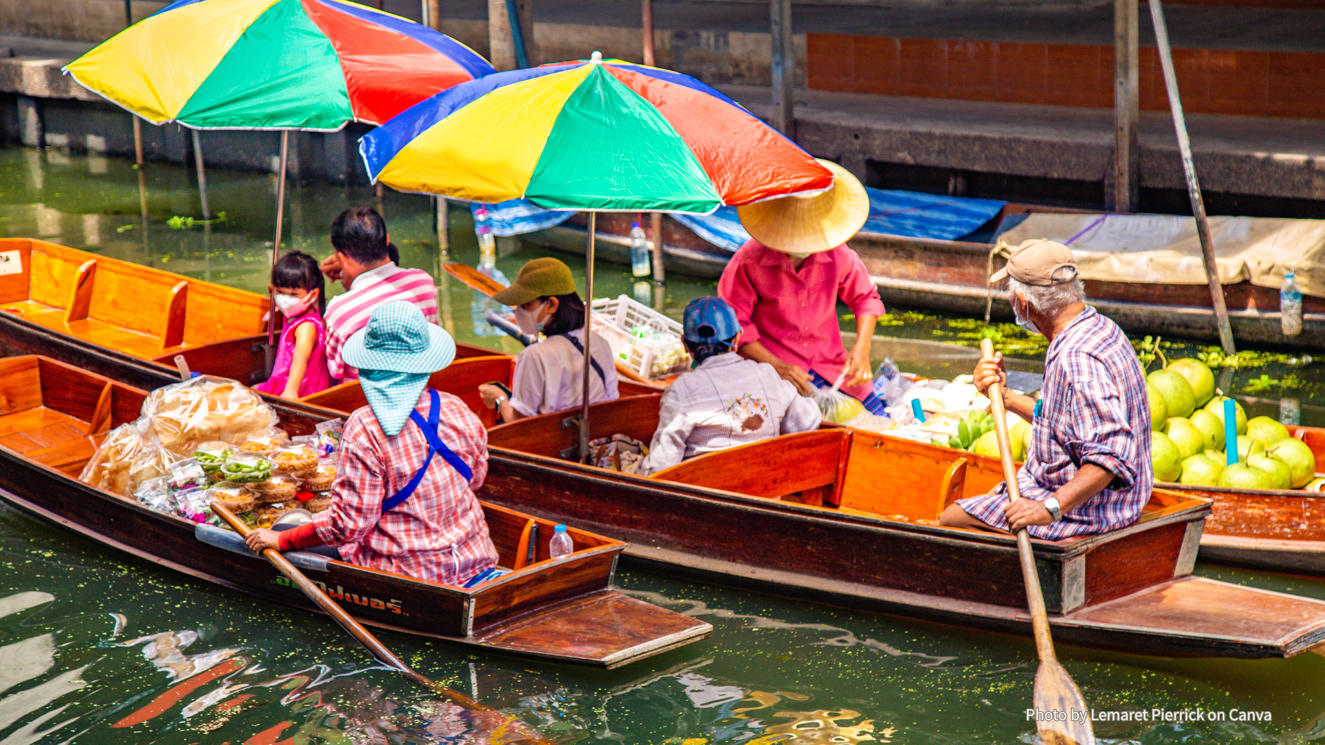 Thai Floating Market, Sabai Sabai Vendors in sun hats paddle produce-laden wooden boats shaded by colorful striped umbrellas at a Thai floating market on a green canal.
